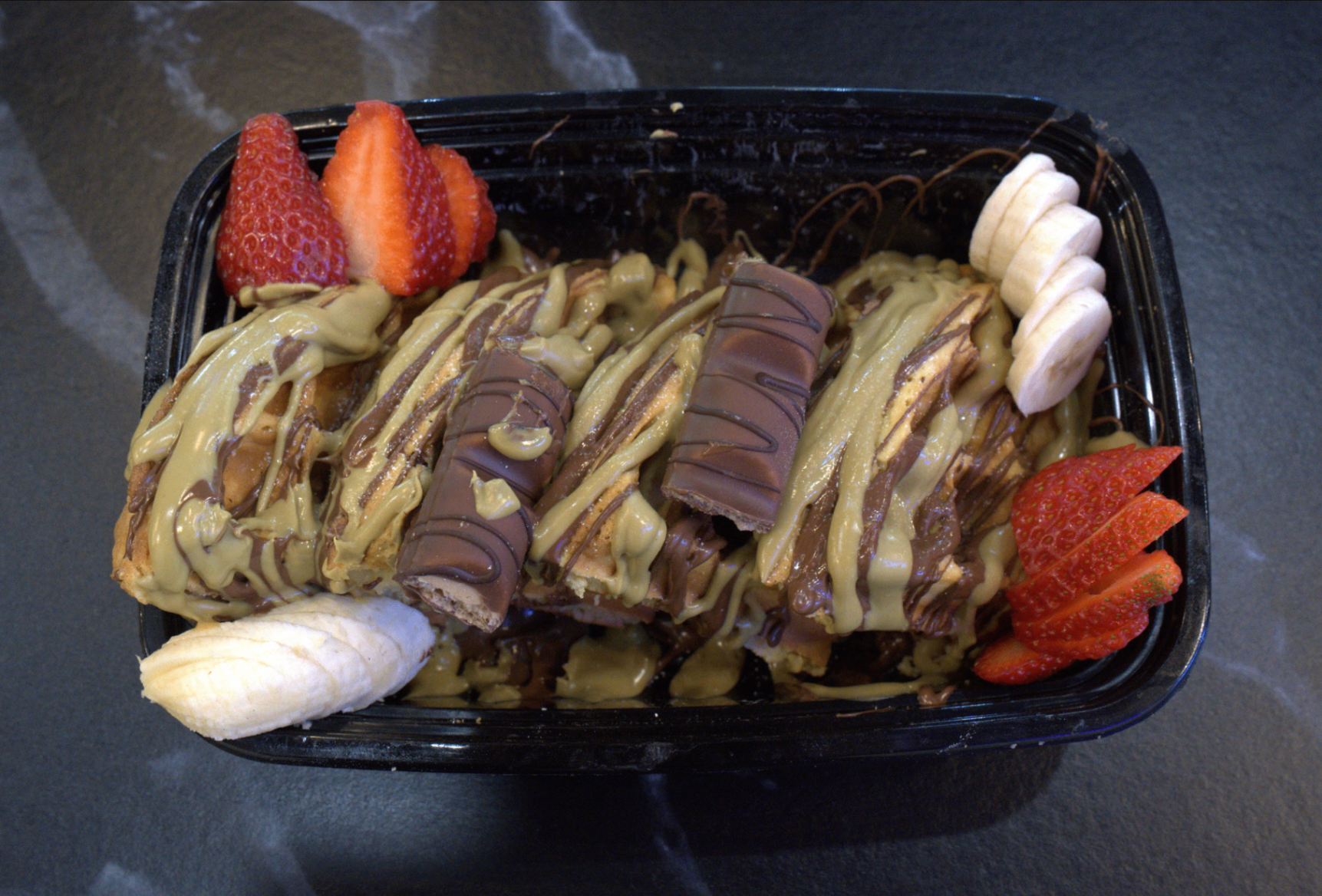 Loaded waffle drizzled with pistachio and Nutella, topped with Kinder Bueno, strawberries and banana