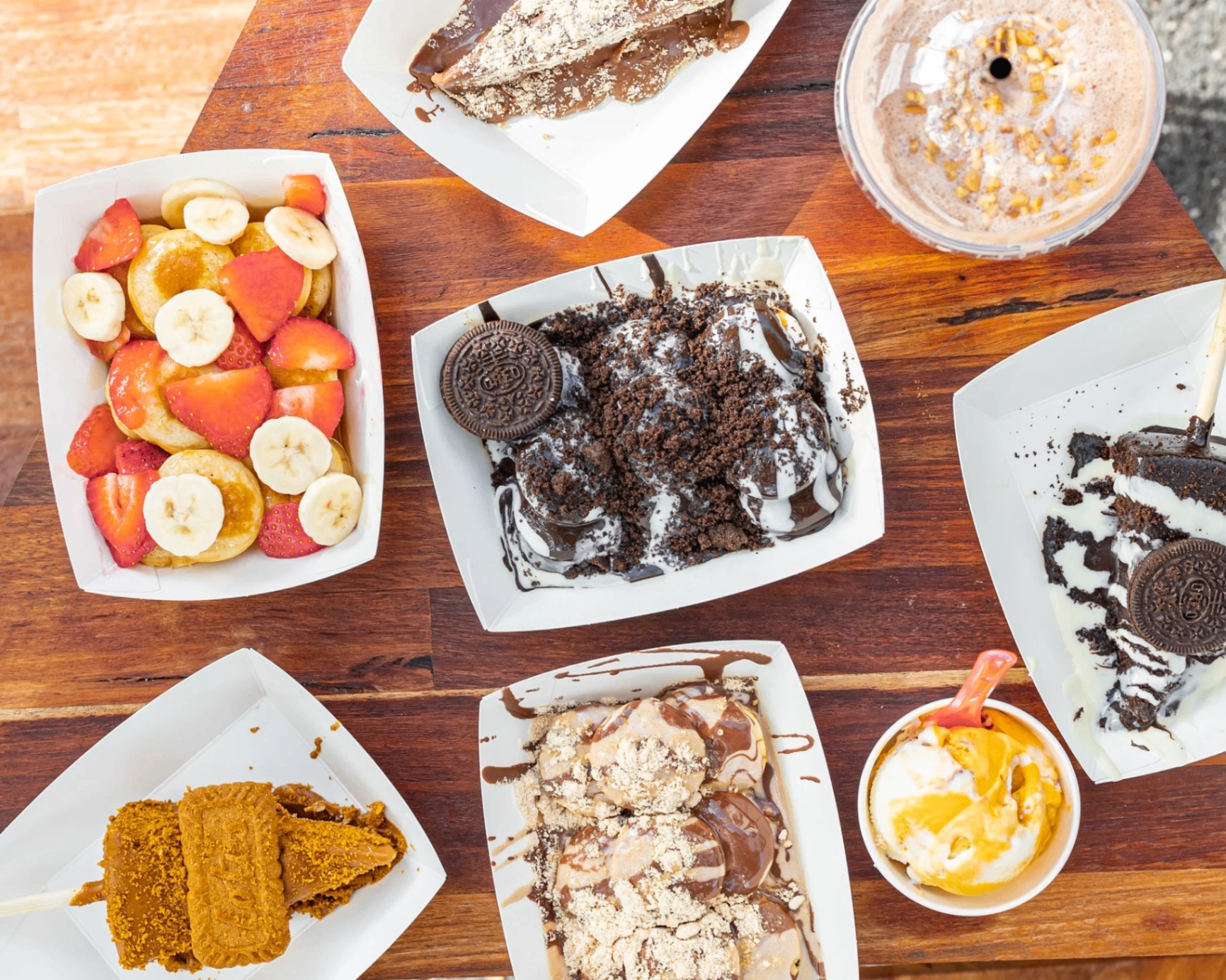 Overhead spread of Crave 'N' Go desserts on a wooden table — strawberry banana pancakes, Oreo loaded cup, Biscoff stick, milkshake and more